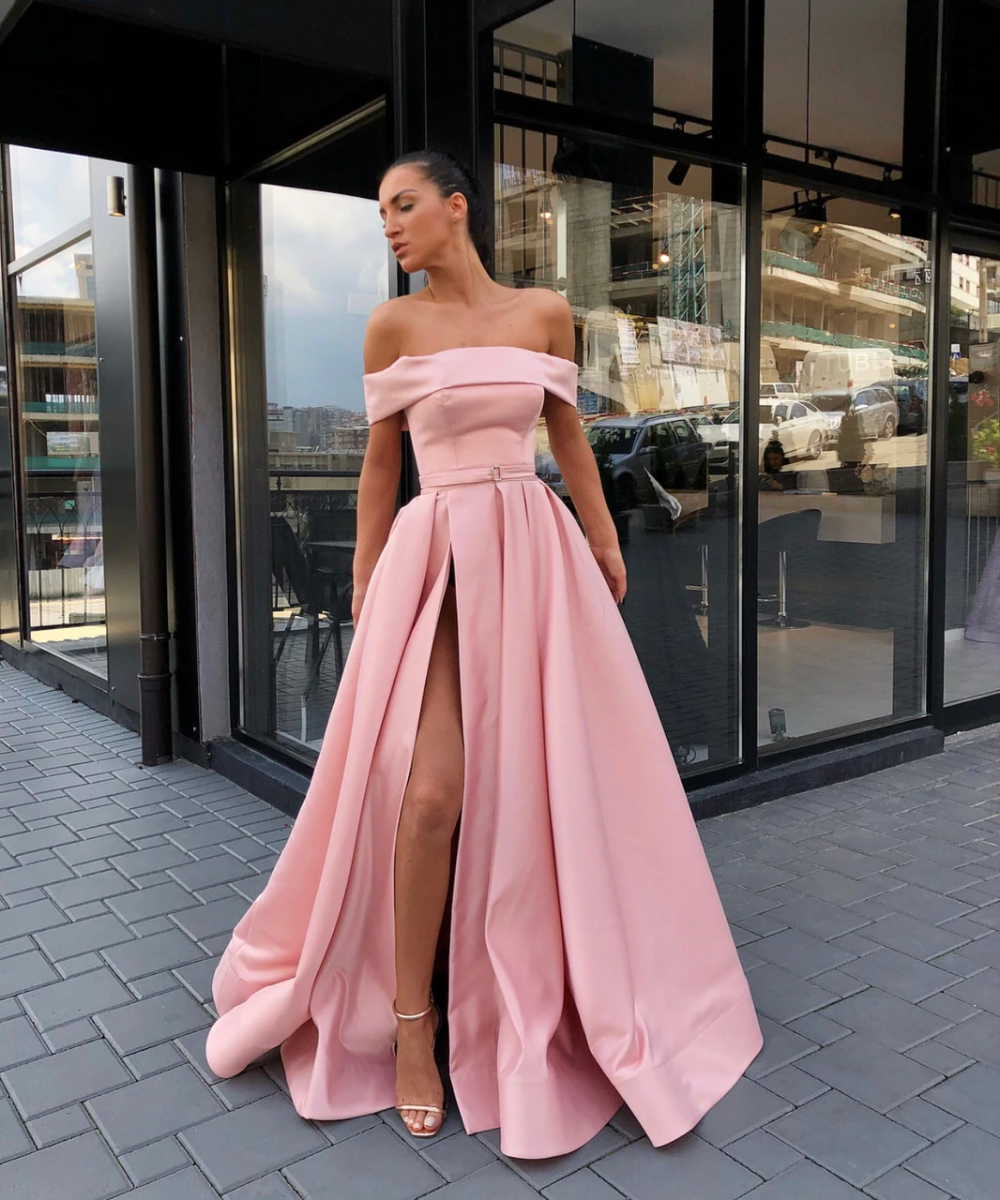 A woman standing in front of a glass door wearing a pink off-the-shoulder a-line prom dress with a satin finish.